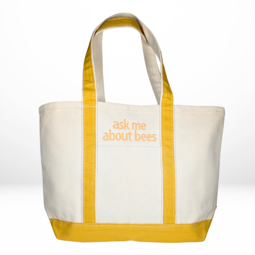 A beige tote bag with yellow handles and base, featuring centered yellow embroidery that reads “ask me about bees.”