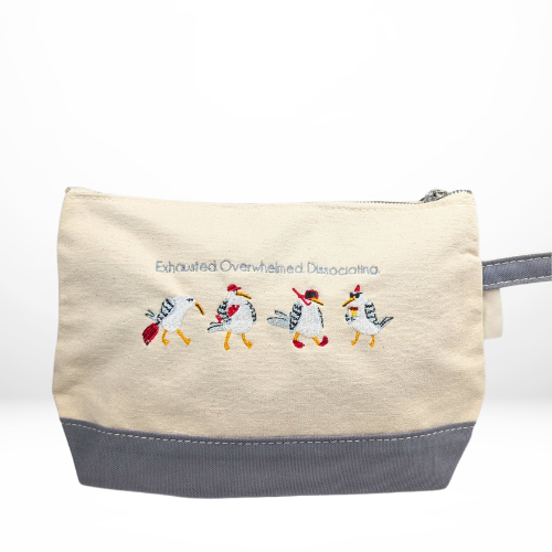 A beige zippered pouch with a gray base, featuring embroidery of four expressive seagulls and the words “Exhausted Overwhelmed Dissociating” stitched above them.