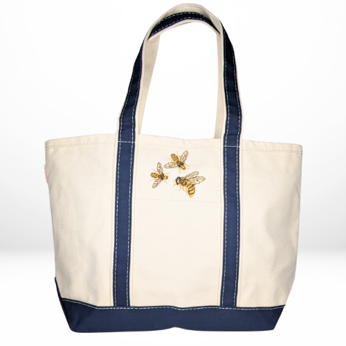 A beige canvas tote with navy blue accents, featuring a trio of golden honey bees embroidered in flight on the front.
