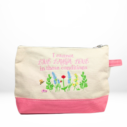 A beige and pink zippered pouch with embroidered text reading, "I cannot LIVE LAUGH LOVE in these conditions," above colorful stitched flowers and a bee.