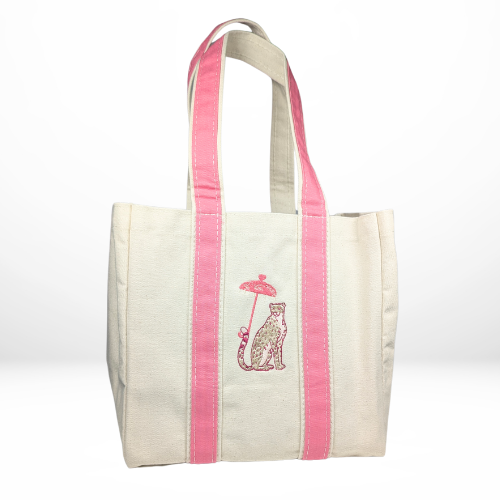 A beige canvas tote bag with coral pink straps, embroidered with a poised leopard sitting beneath a decorative parasol.